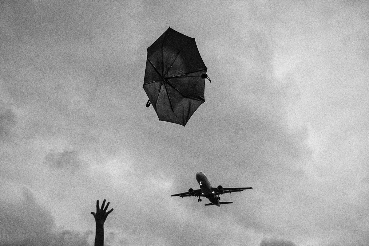 Black-and-white, looking up: a black umbrella tumbling loose and inverted against a grey sky, a bare hand thrust up from the bottom edge reaching for it, and an airliner on final approach below and behind